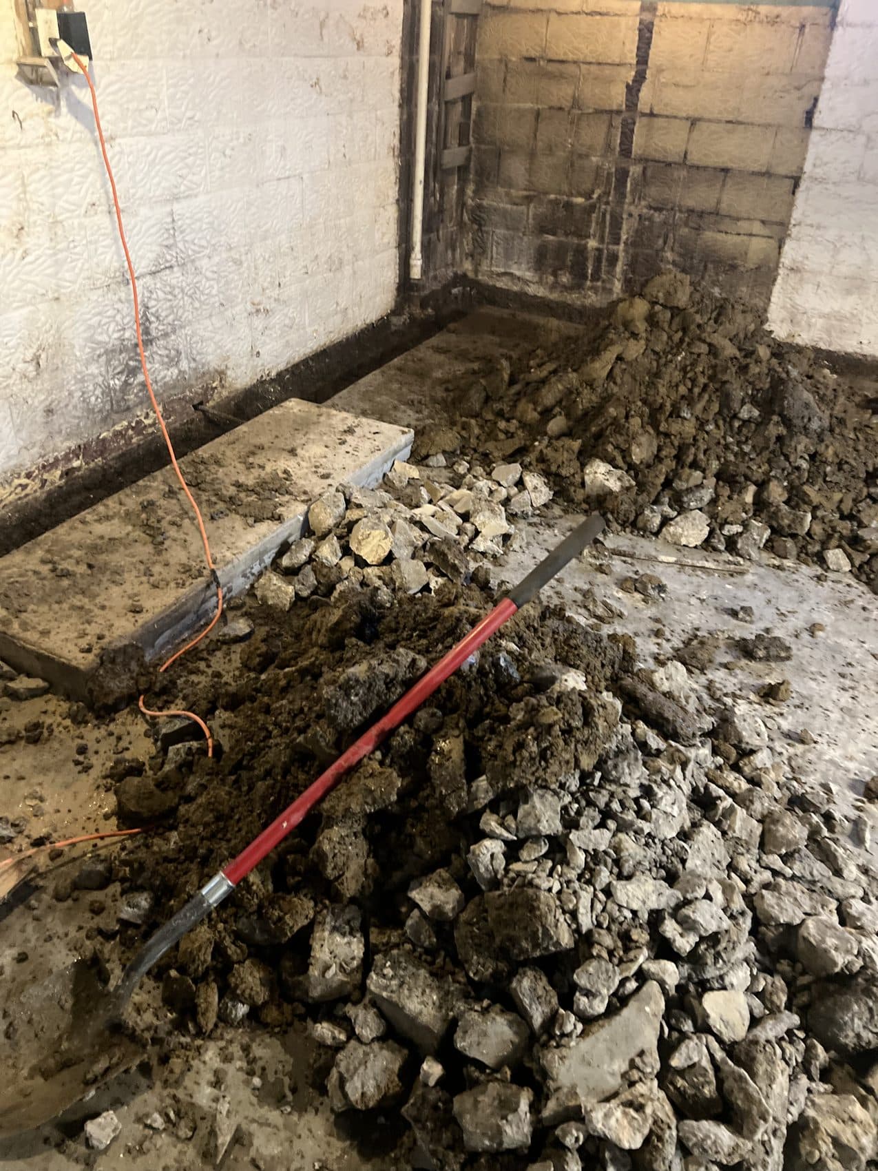 A basement floor opened up along a damp block wall, ready for a new drainage system