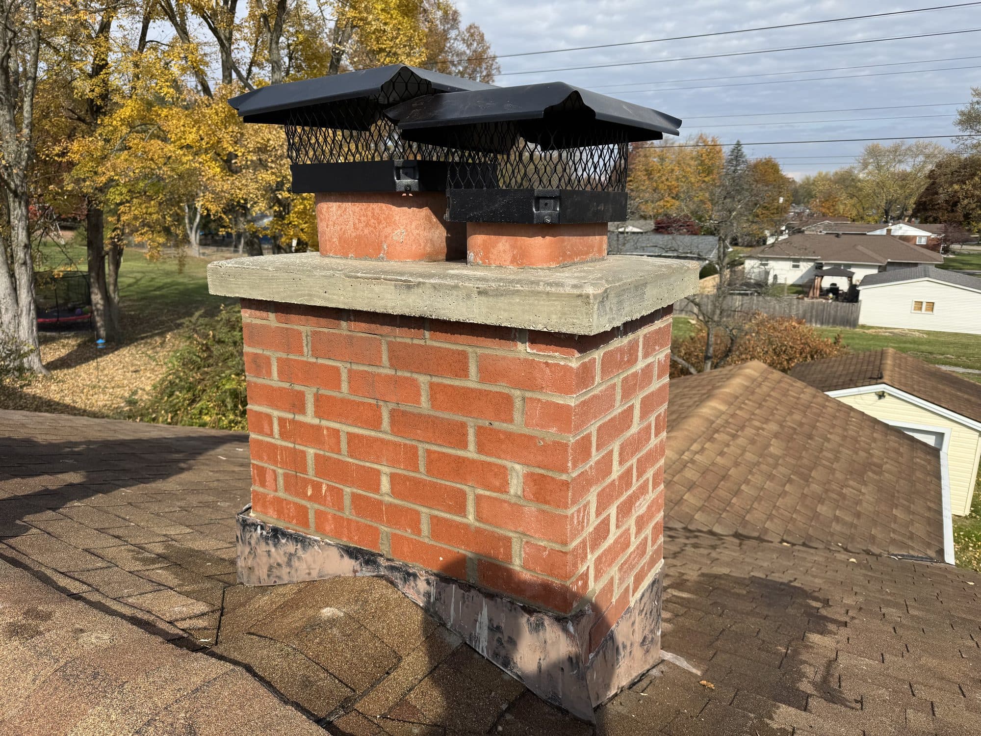 Twin-flue red brick chimney rebuilt with a fresh crown and two caps over a fall neighborhood
