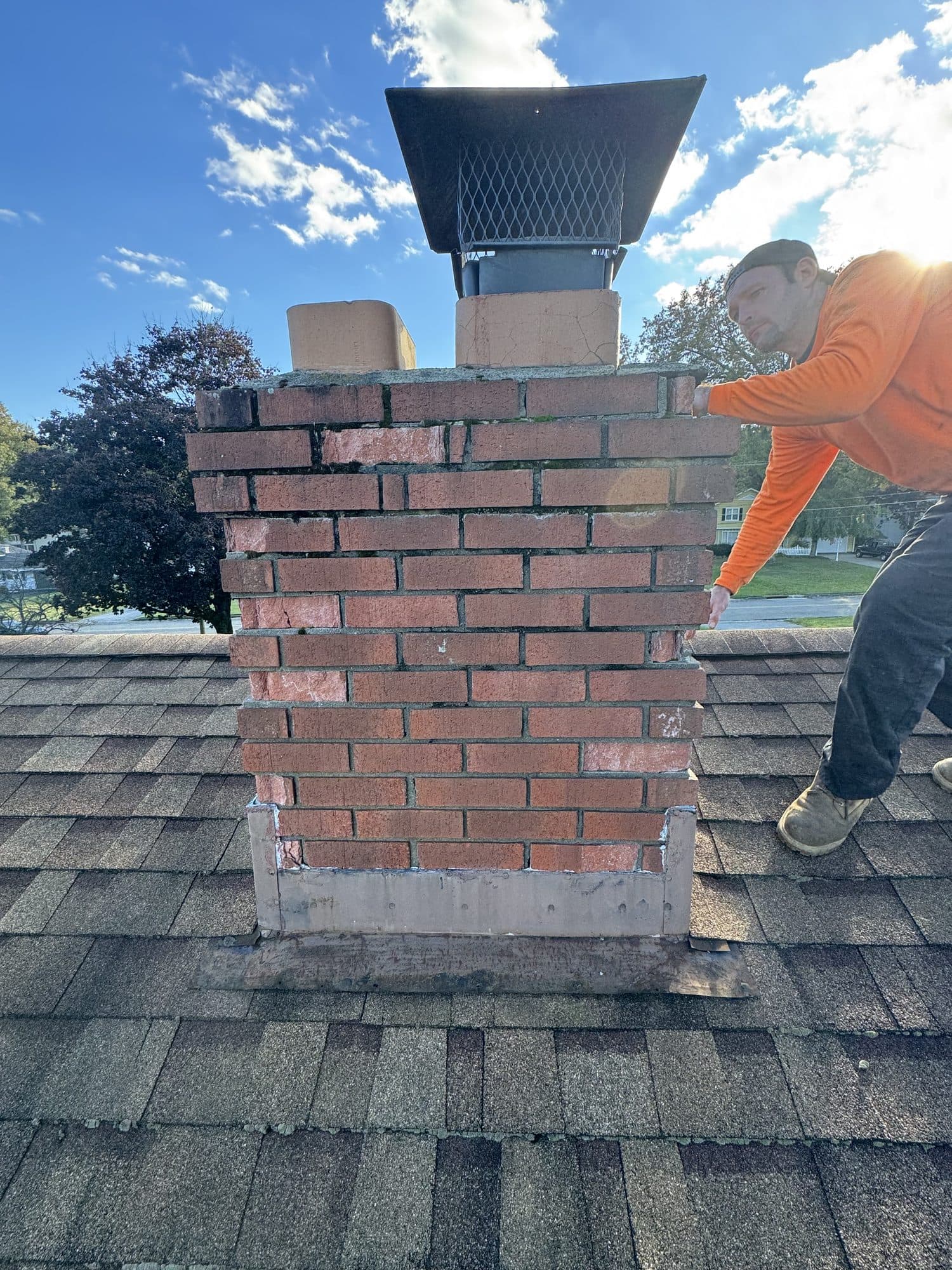 A HydroGuard mason on a roof setting the cap on a freshly rebuilt brick chimney, sun breaking behind